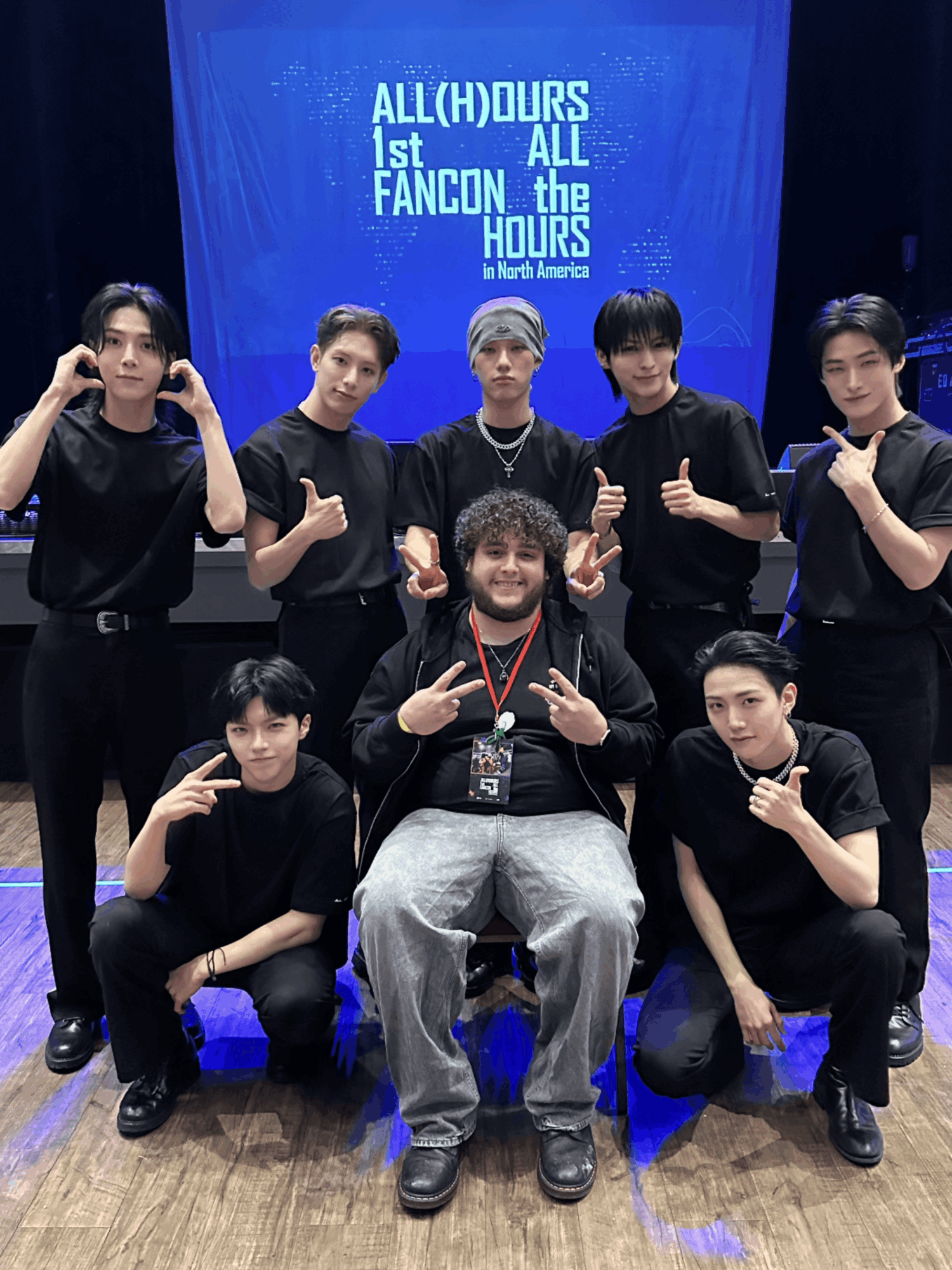 Group of seven people, six standing and one sitting, posing in front of a blue screen with text, all wearing black clothing.