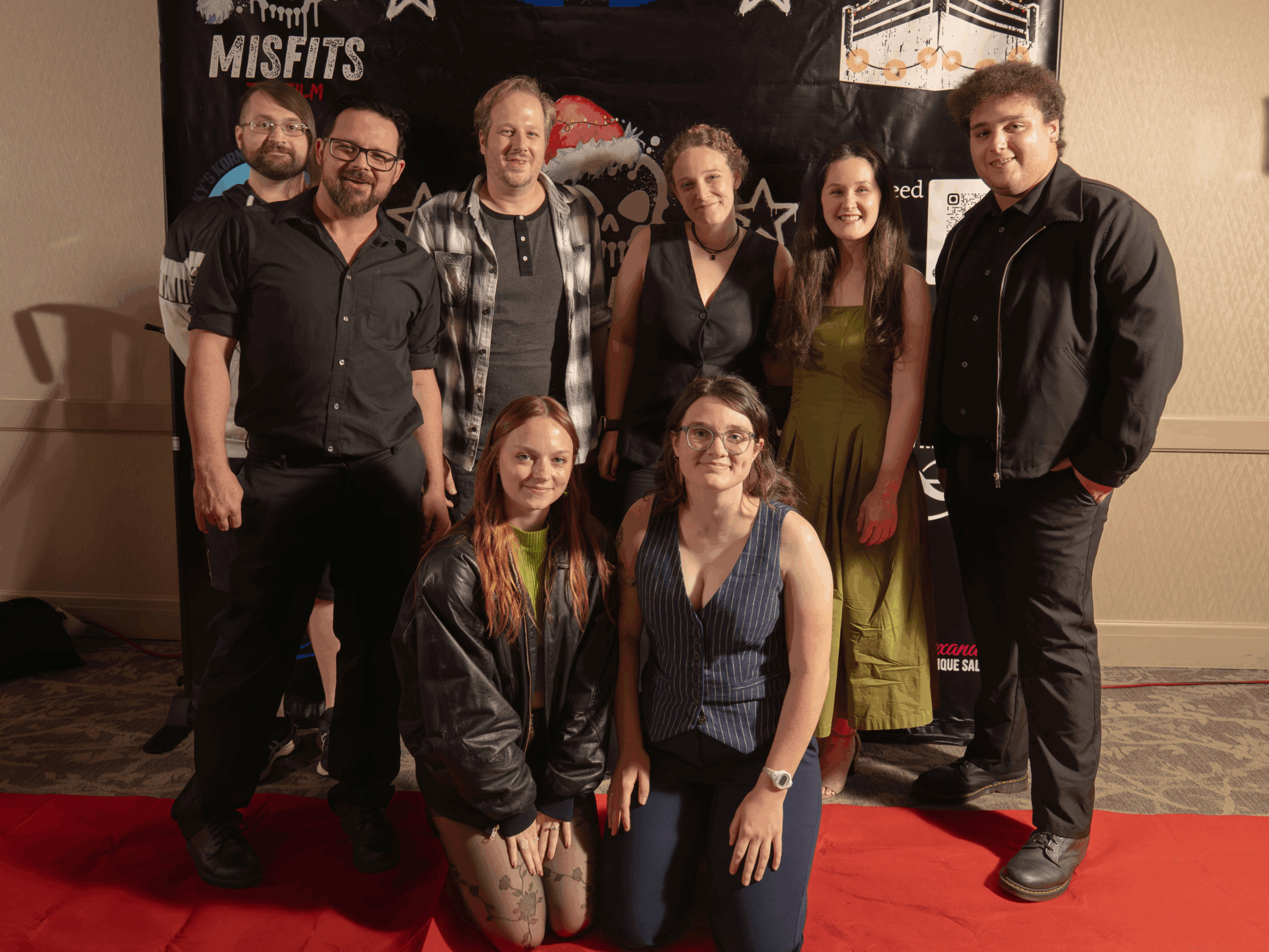Group of eight people standing on a red carpet in front of a black backdrop with band logos, smiling at the camera.