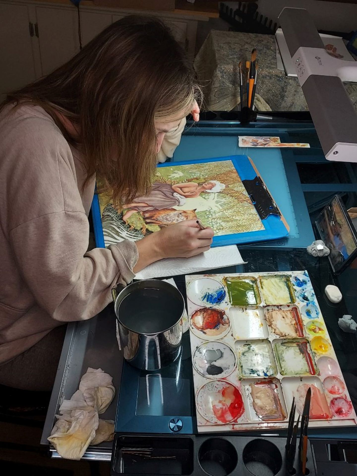 Person painting on canvas with watercolor paints, workspace with brushes, palette, and art supplies, indoor setting.