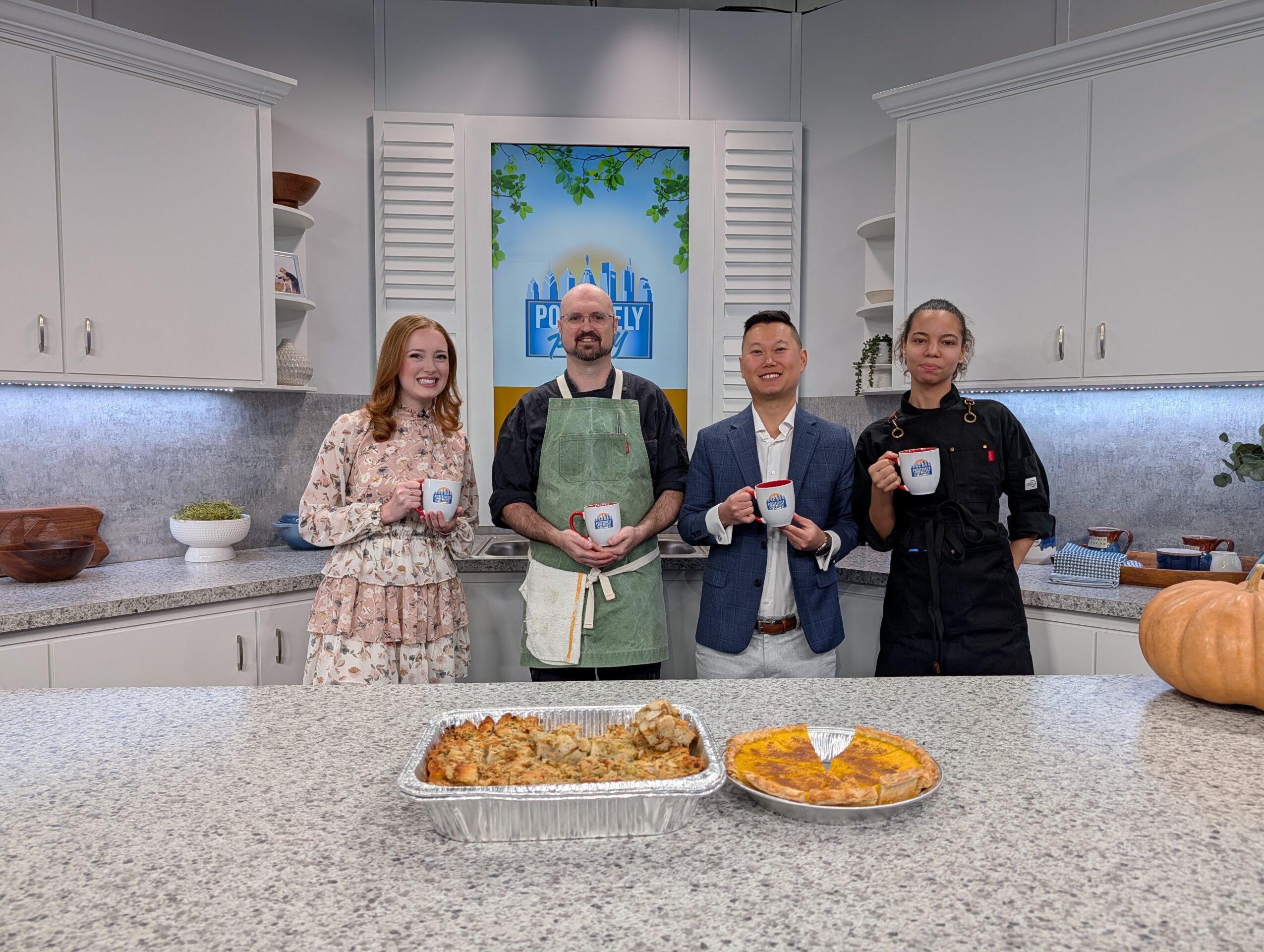 Four people stand behind a kitchen counter with baked goods, smiling, in a bright kitchen with white cabinets and a colorful painting.