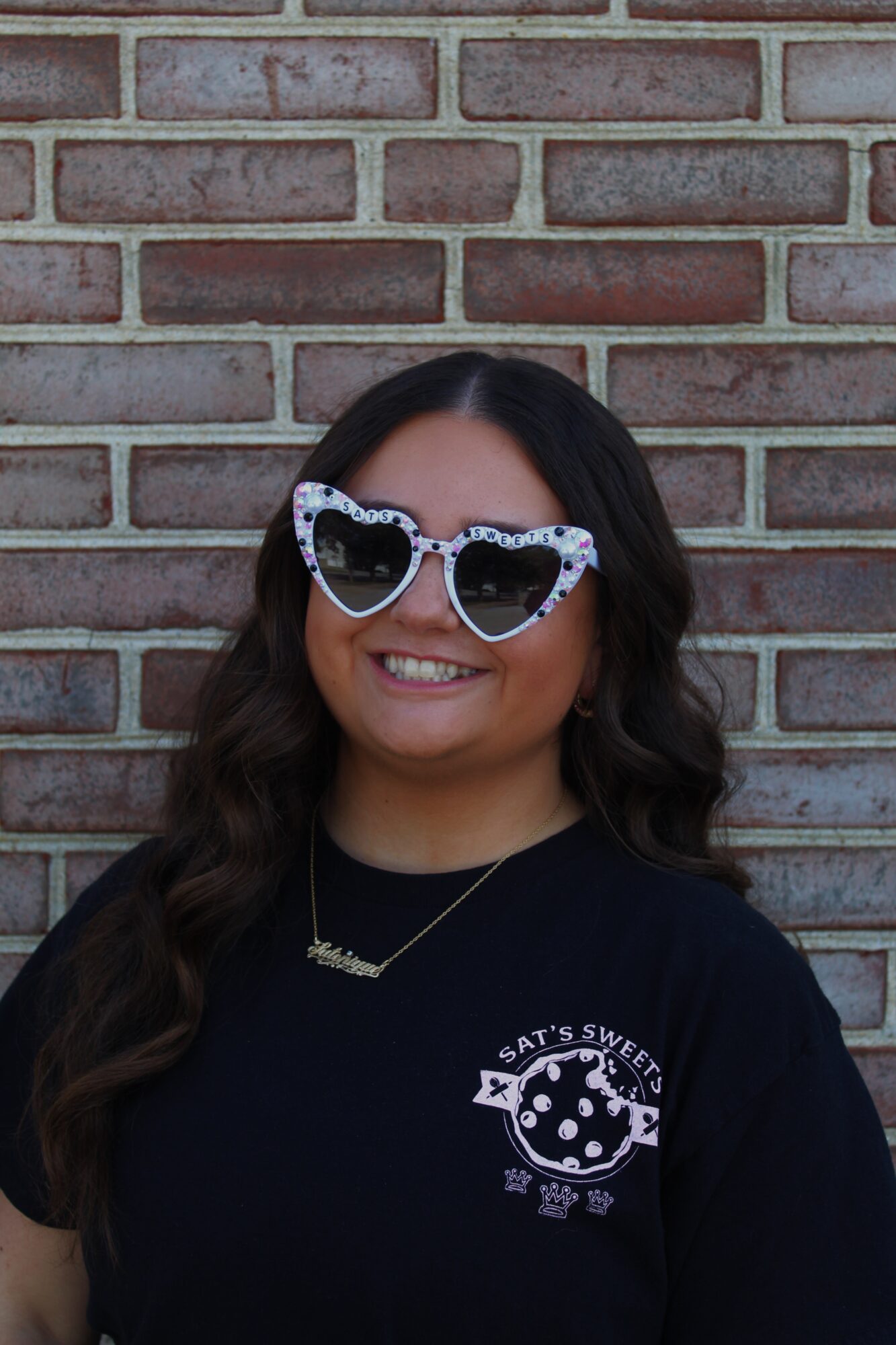 Person with long dark hair wearing heart-shaped sunglasses and a black shirt standing in front of a brick wall.
