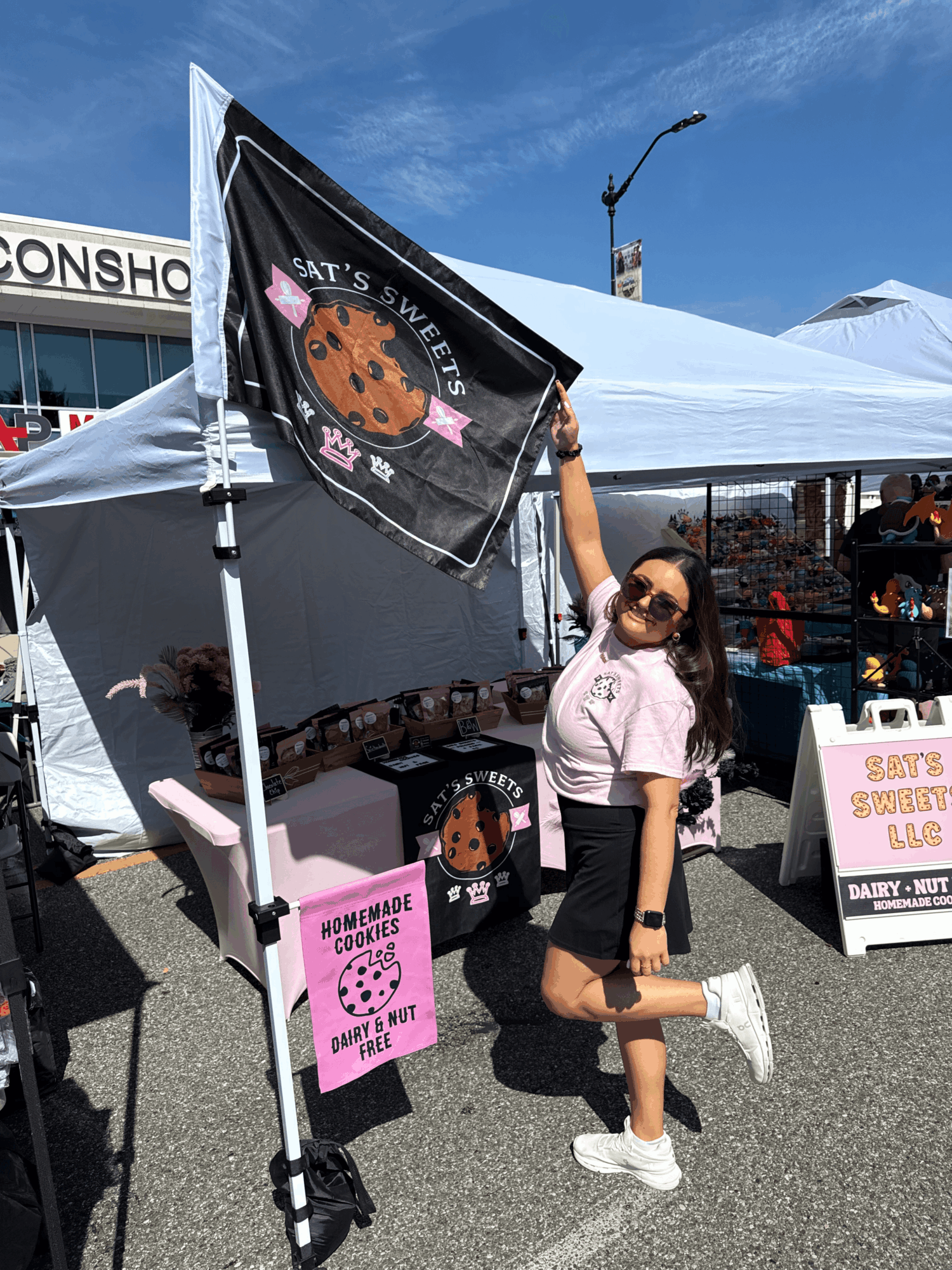 Young woman with sunglasses and a pink shirt, standing outdoors under a tent, holding a flag with a cookie design, smiling.