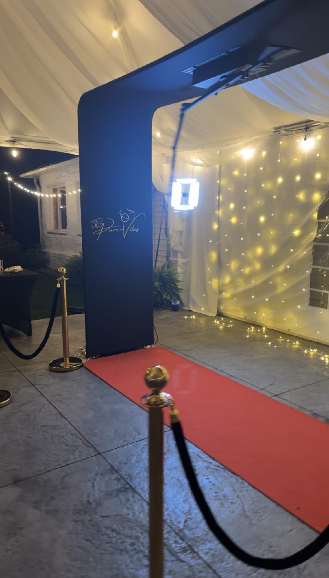 Red carpet with black stanchions and velvet ropes leading to a decorated entrance with string lights and a sign, indoors.