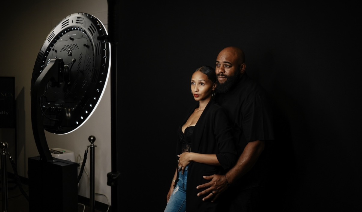 Two people stand close together in a dark room, with a large clock on the left side of the image.