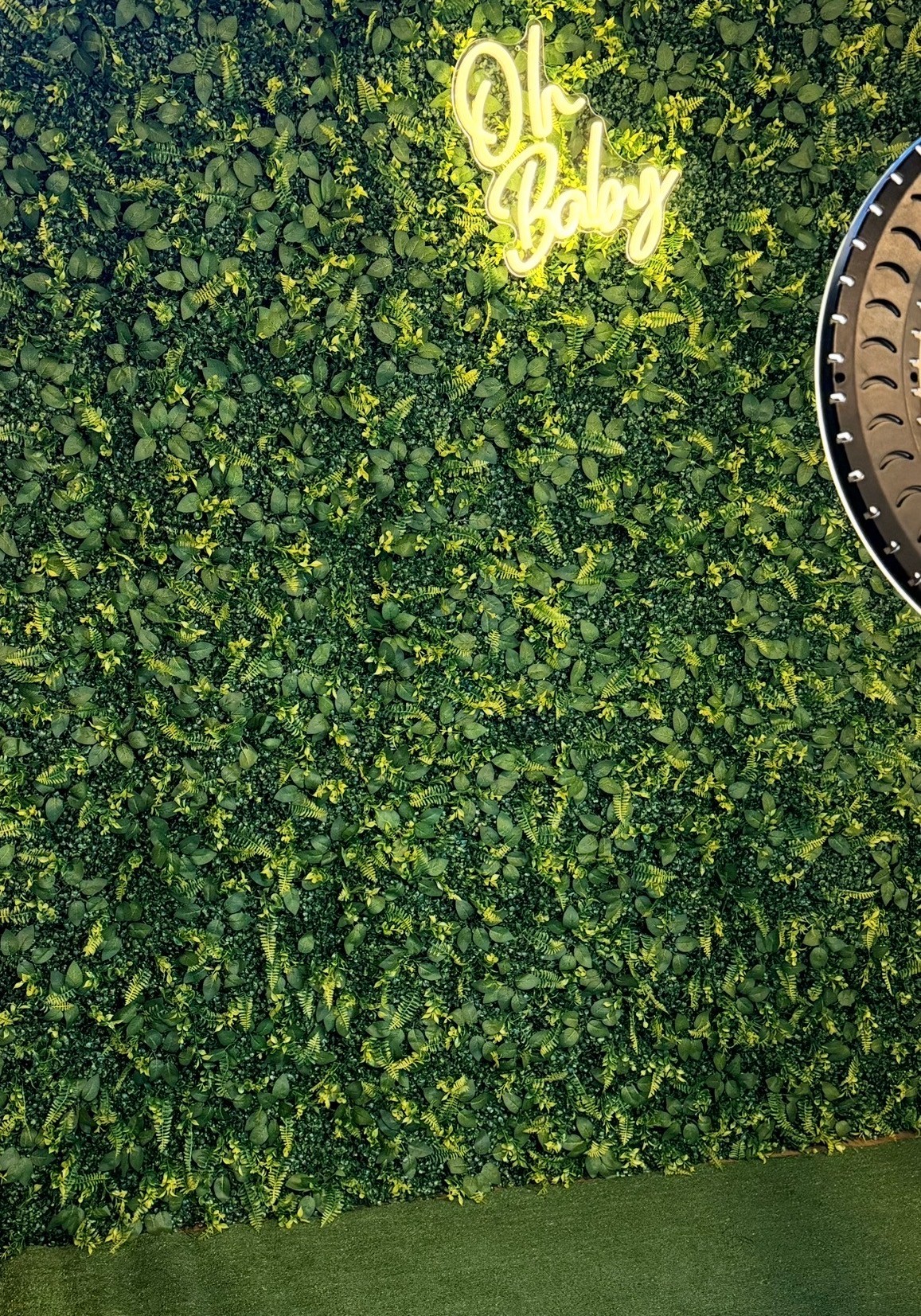 Green leafy wall with a yellow neon sign reading 'Oh Love' and part of a round clock visible on the right.
