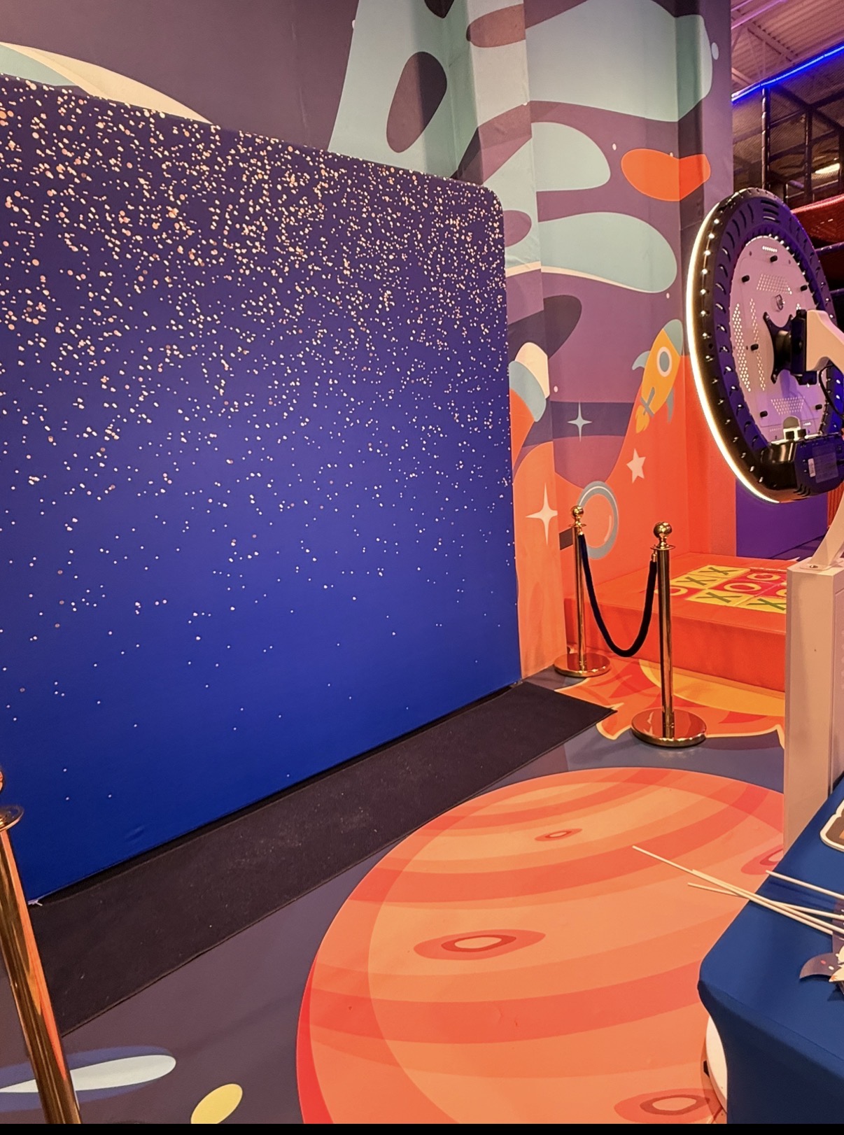 Colorful indoor space with a large starry backdrop, abstract wall art, and a decorative clock, with a red carpet and velvet rope barrier.