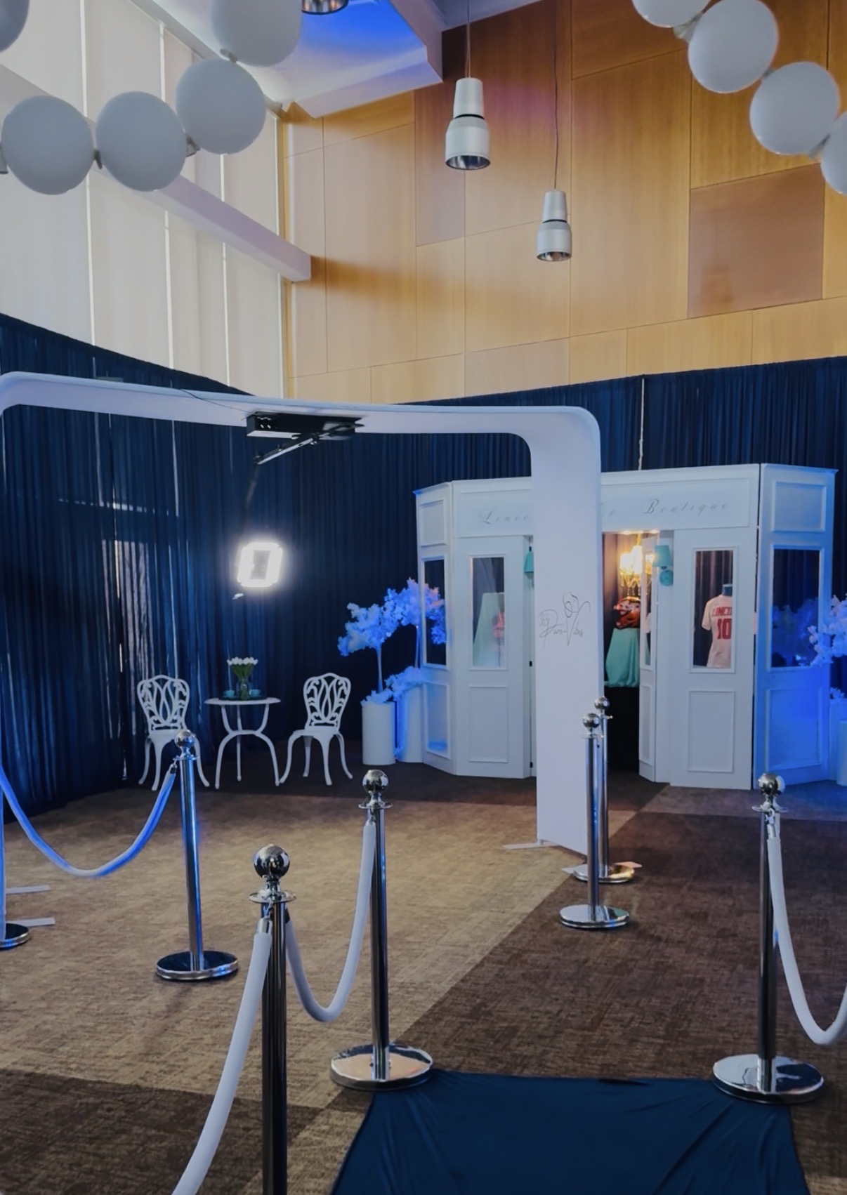 Indoor space with white booths, chairs, and blue lighting, decorated with white balloons and a brown carpeted area.