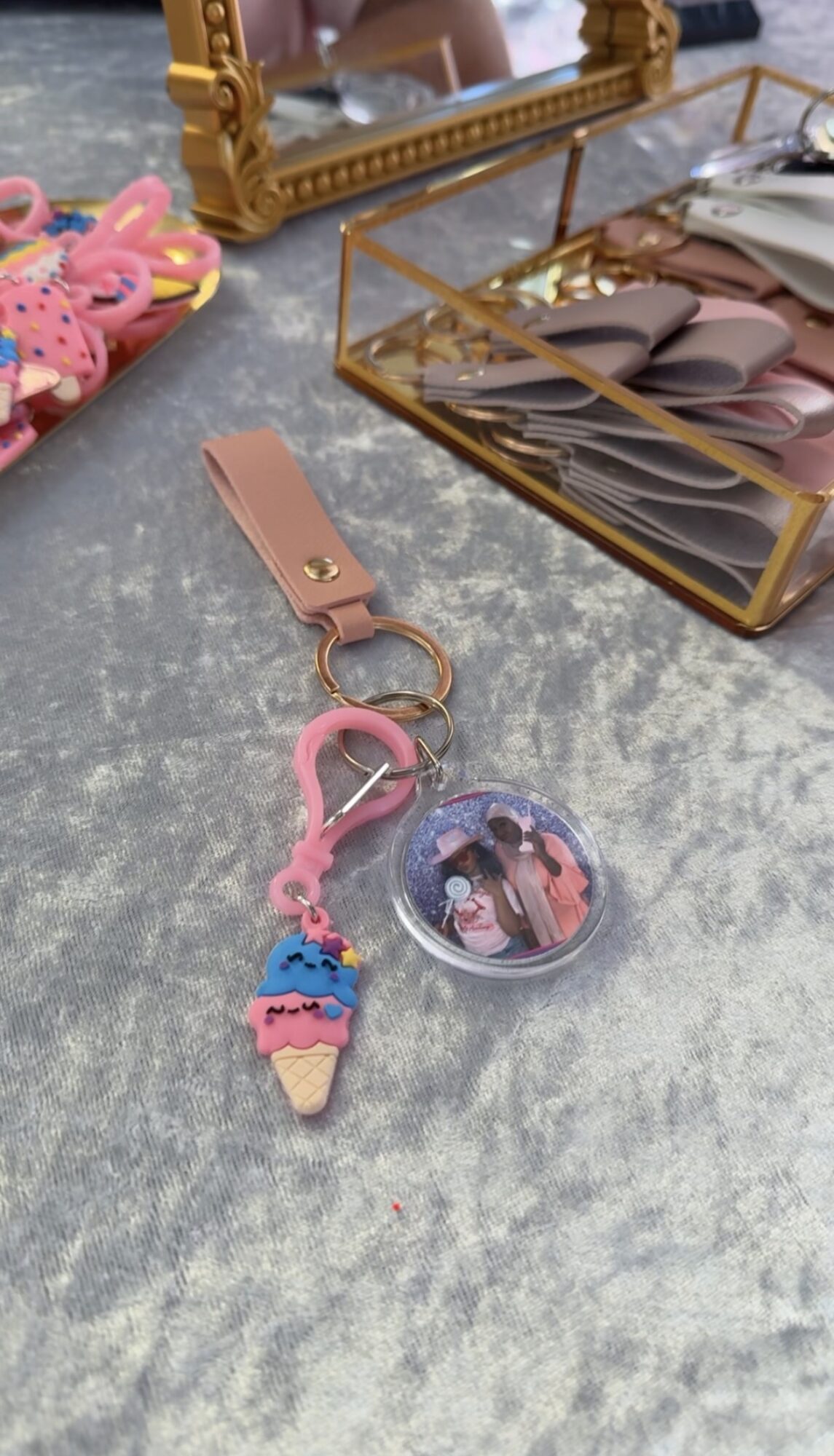 Keychain with pink ice cream cone and a clear round charm, on a gray surface with jewelry and accessories nearby.