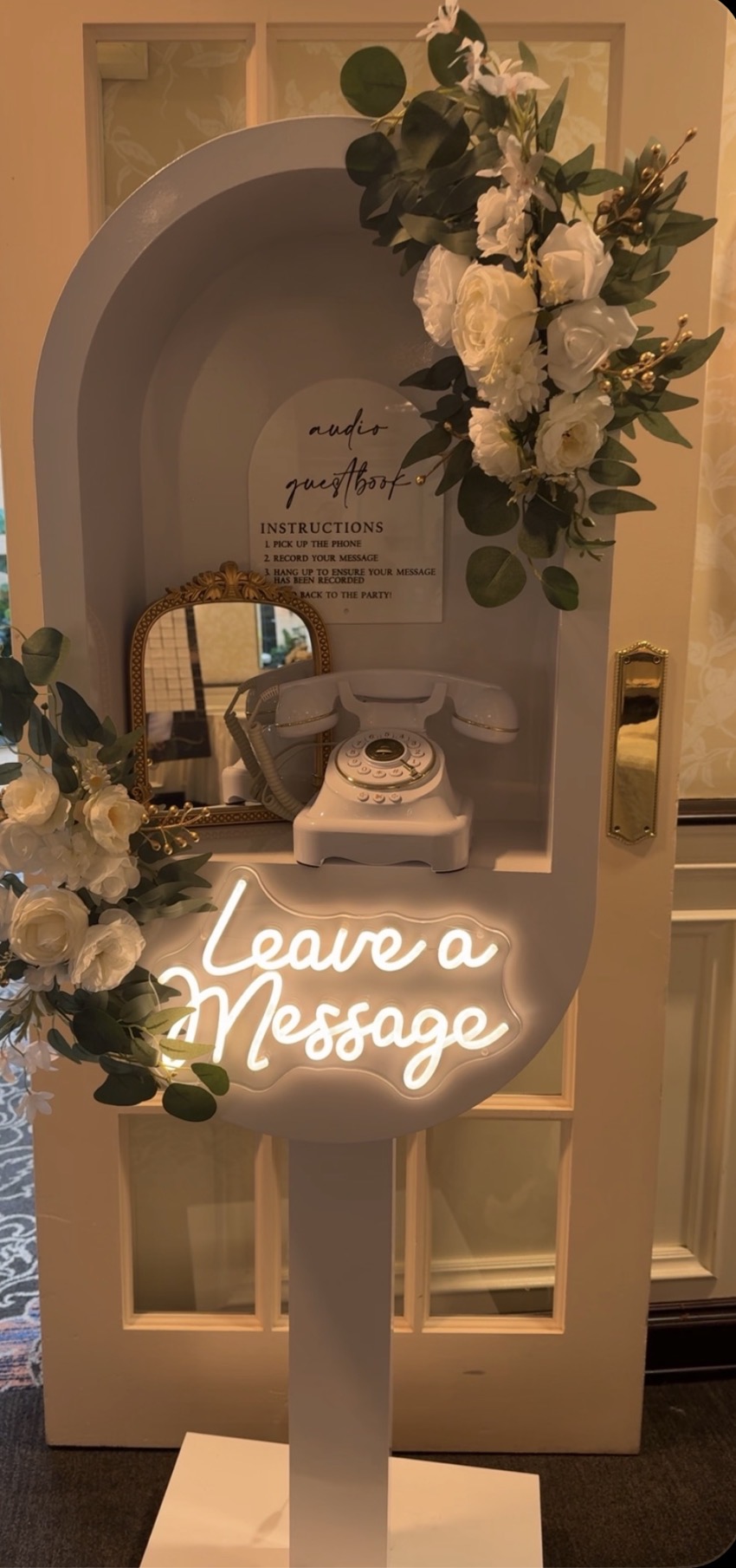 Decorative display with white flowers, a small mirror, a phone, and a neon sign reading 'Leave a Message'.