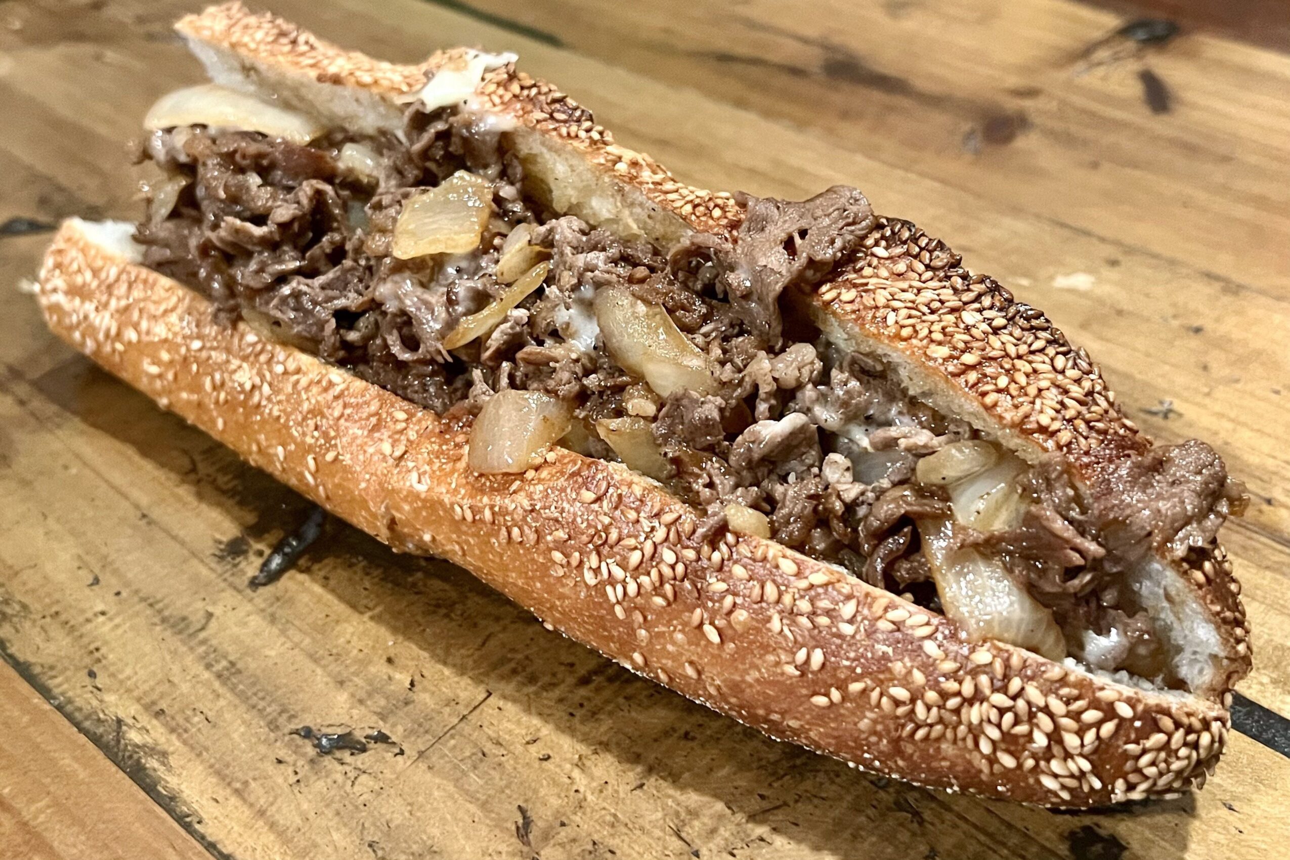 Sandwich with beef, onions, and sesame seed bun on wooden surface.