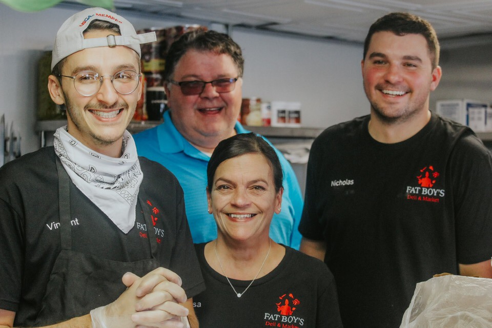 Four smiling people in a kitchen, two wearing black shirts with red logos, one in a blue shirt, and one in a white cap and glasses.