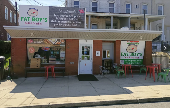 storefront with signs for Fat Boy's deli and market, outdoor seating with red and green chairs, sidewalk in front