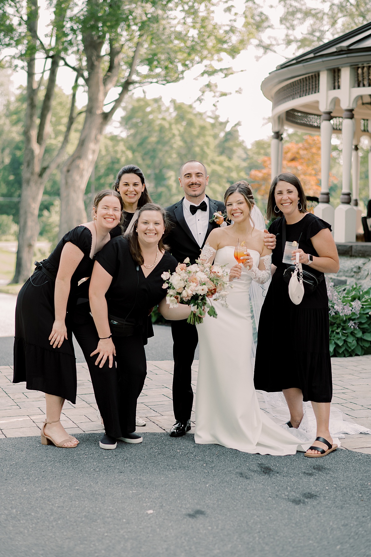 Group of six people, including a bride in a white wedding dress holding a bouquet, standing outdoors near a gazebo, smiling.
