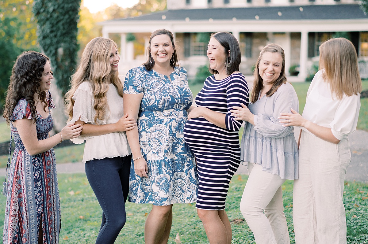 Six women standing outdoors, smiling and talking, with a house and trees in the background.