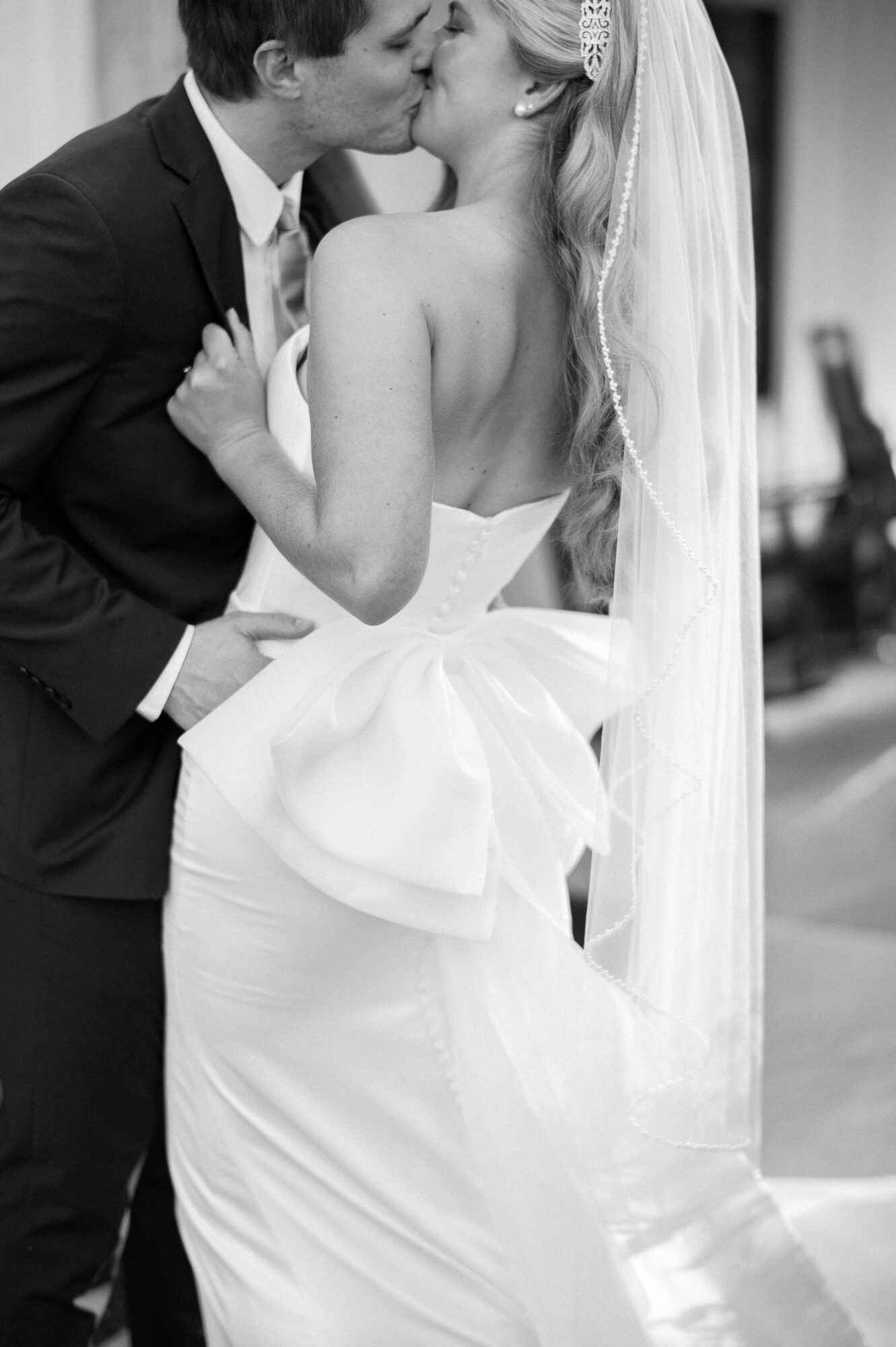 A bride and groom kiss, bride in a strapless gown with a large bow, groom in a suit, wedding setting.
