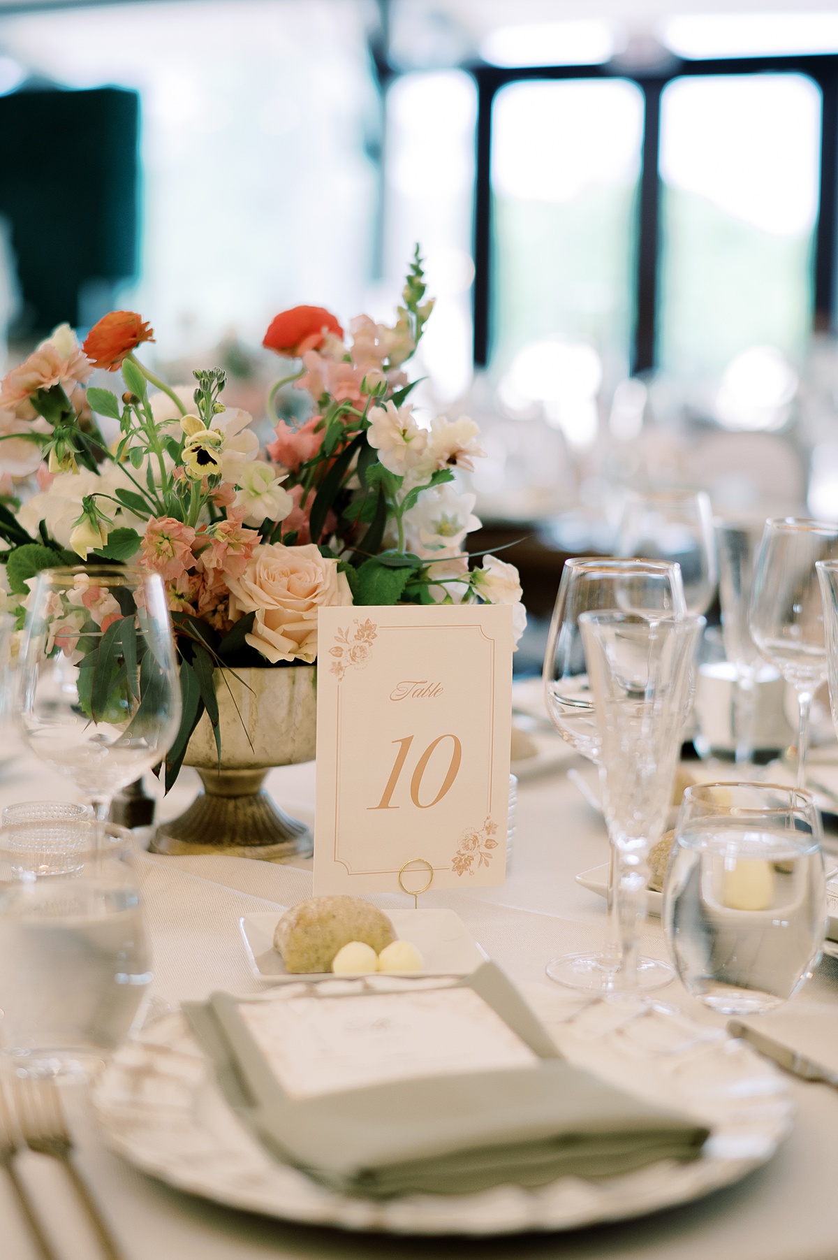Elegant table setting with a floral centerpiece, table number card, glasses, and napkins, in a bright room with large windows.