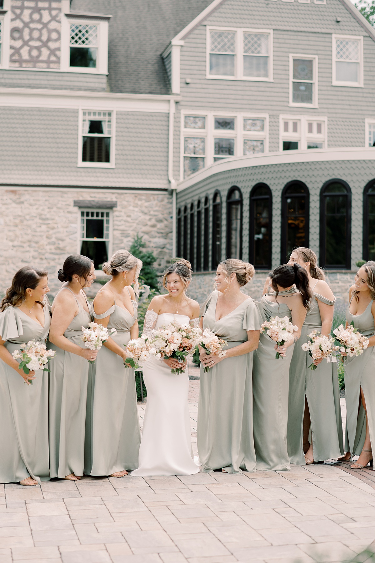 Group of women in light-colored dresses holding bouquets, standing outdoors in front of a large building with multiple windows.