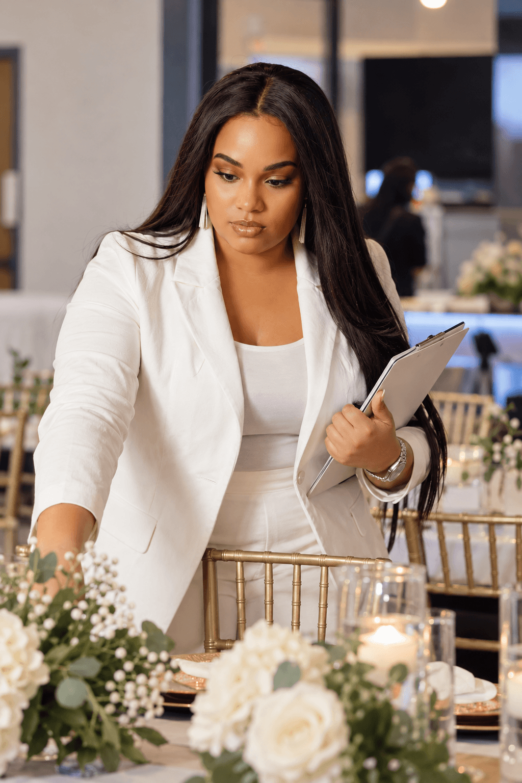Woman in white blazer holding a tablet, arranging flowers at a table, in a well-lit indoor setting.