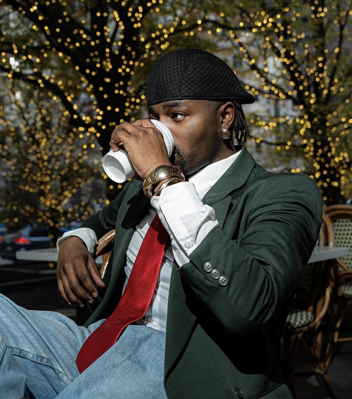 Man in a suit and red tie drinks from a cup outdoors at night with illuminated trees in the background.
