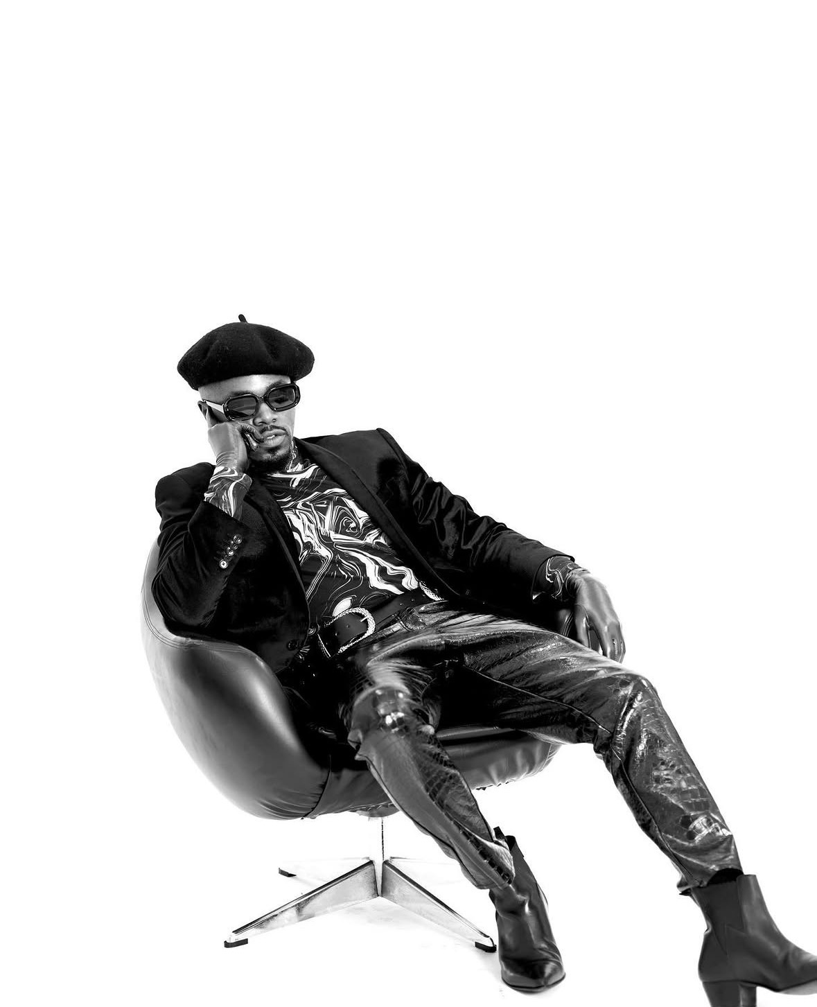 Person sitting in a modern chair, wearing sunglasses, a hat, and stylish clothing, with a relaxed pose.