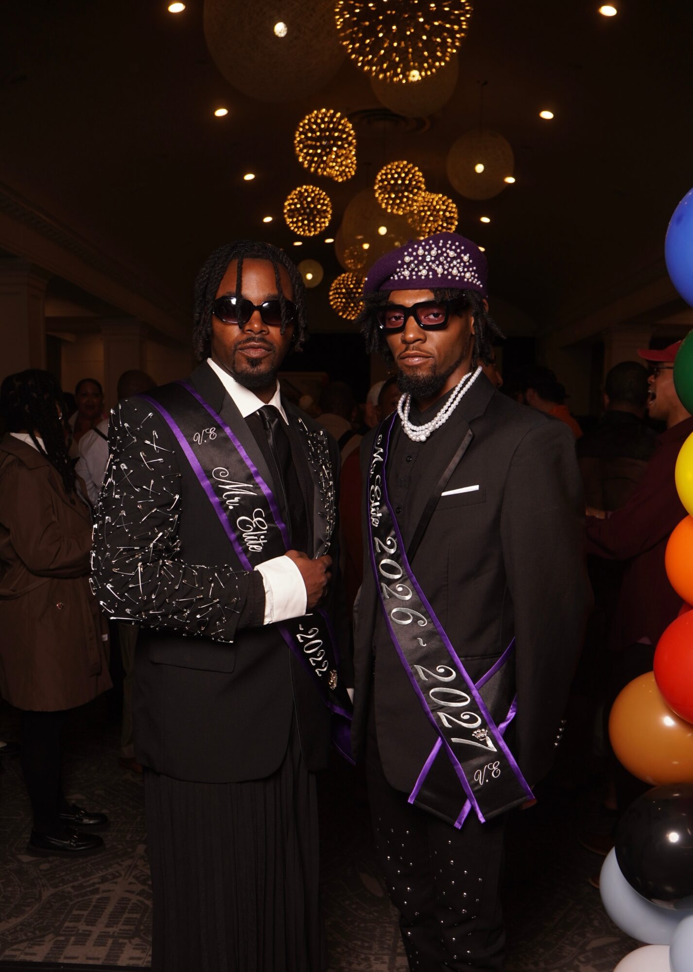 Two men in formal suits and sunglasses pose at an indoor event with hanging spherical lights and colorful balloons.