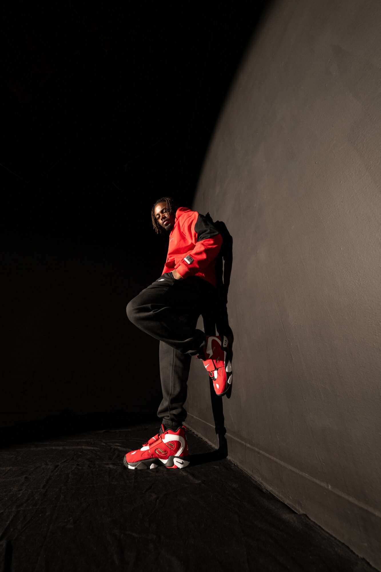 Person in red jacket and black pants leaning against a dark wall with roller skates, in a dimly lit space.