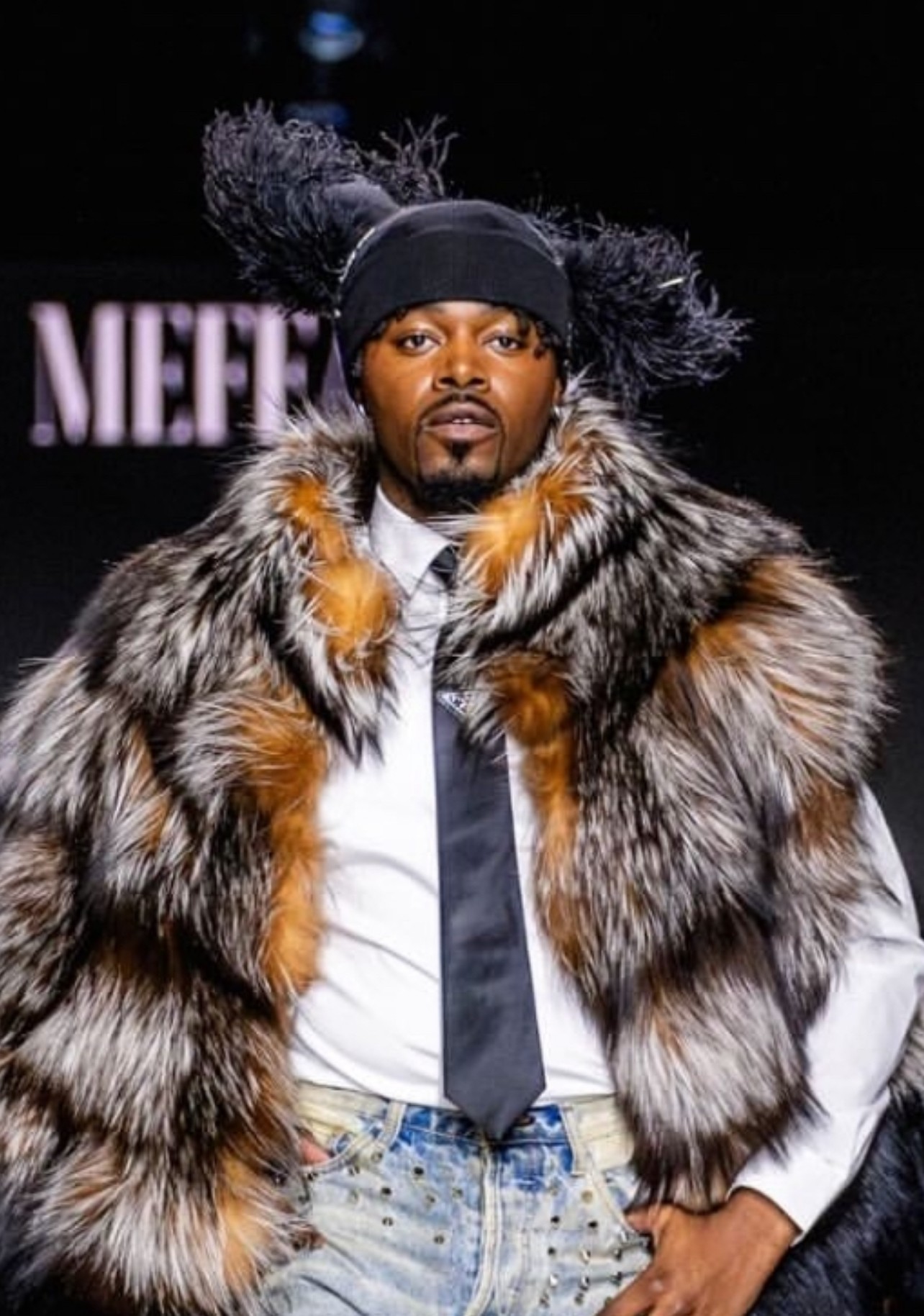 Man on runway wearing a fur coat, black headband, and white shirt with a black tie, with a blurred background.