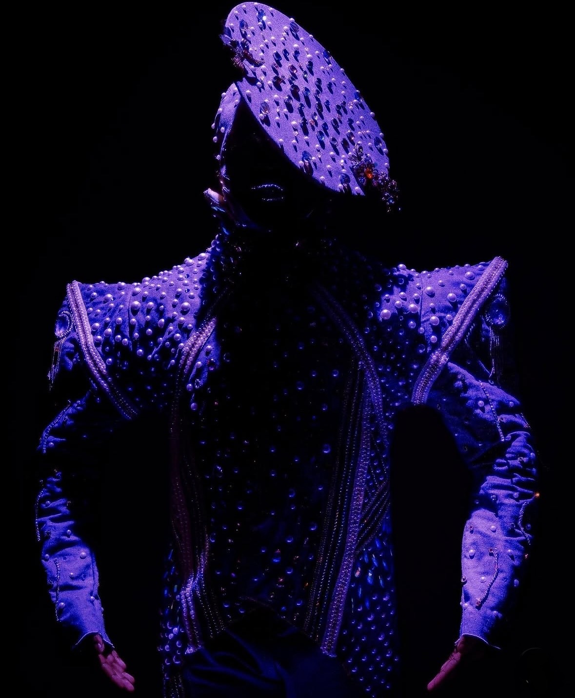 Person in a sparkling, embellished costume with a large, decorated headpiece, illuminated against a dark background.